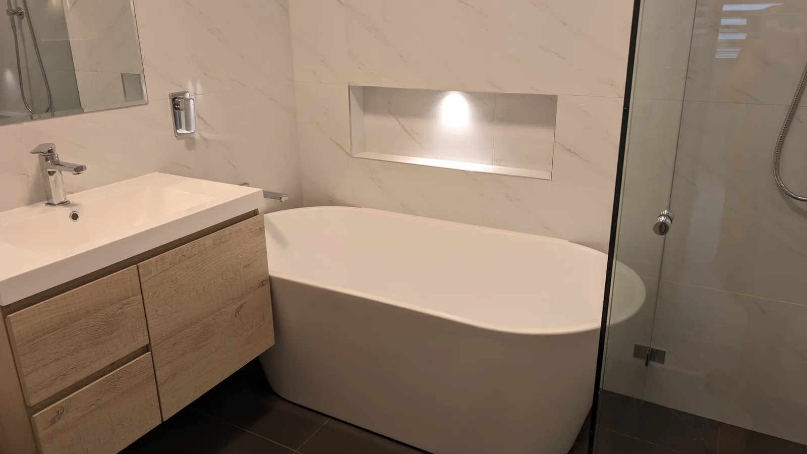 Light-toned bathroom with a timber-look wall-hung vanity, freestanding bath, illuminated wall niche and frameless shower screen.