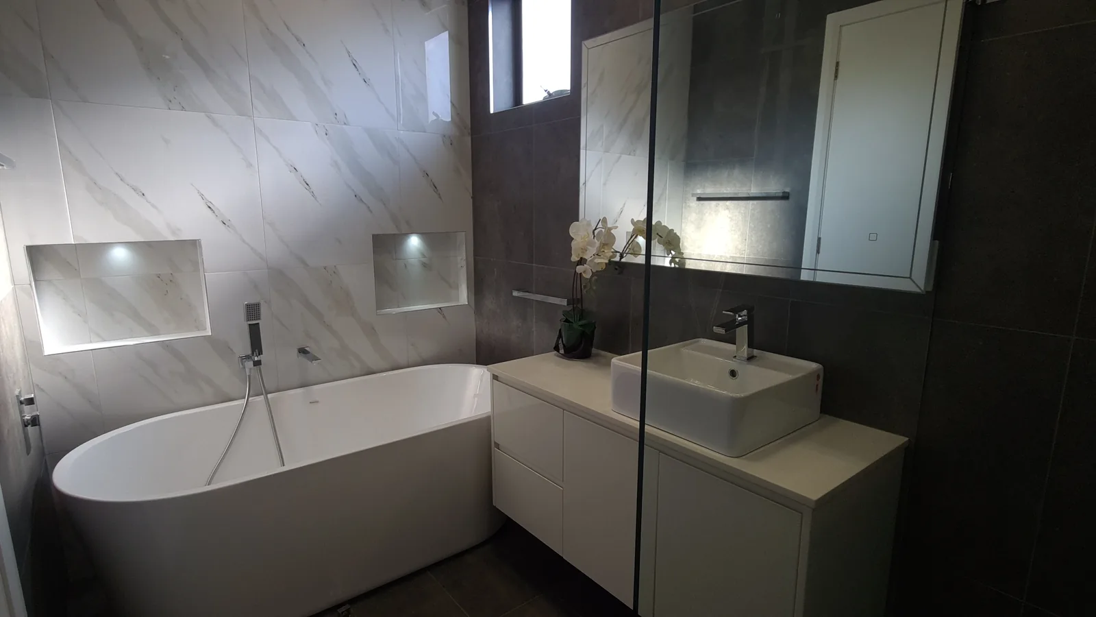 Bathroom with a freestanding bath, wall-hung vanity, dark floor-to-ceiling tiling and two illuminated wall niches.