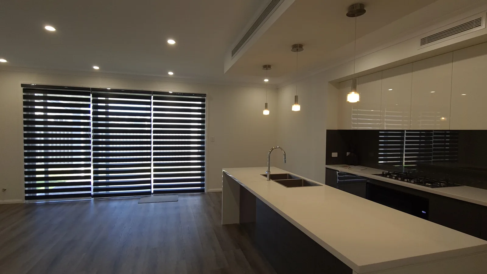 Open-plan kitchen with a long island, double-bowl sink, gloss cabinetry, gas cooktop, pendant lights and wide zebra blinds.
