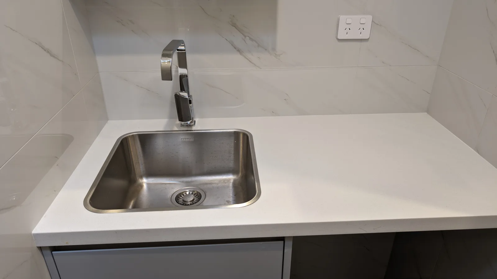 Stainless-steel laundry sink with a chrome mixer set into a white benchtop with a tiled splashback.